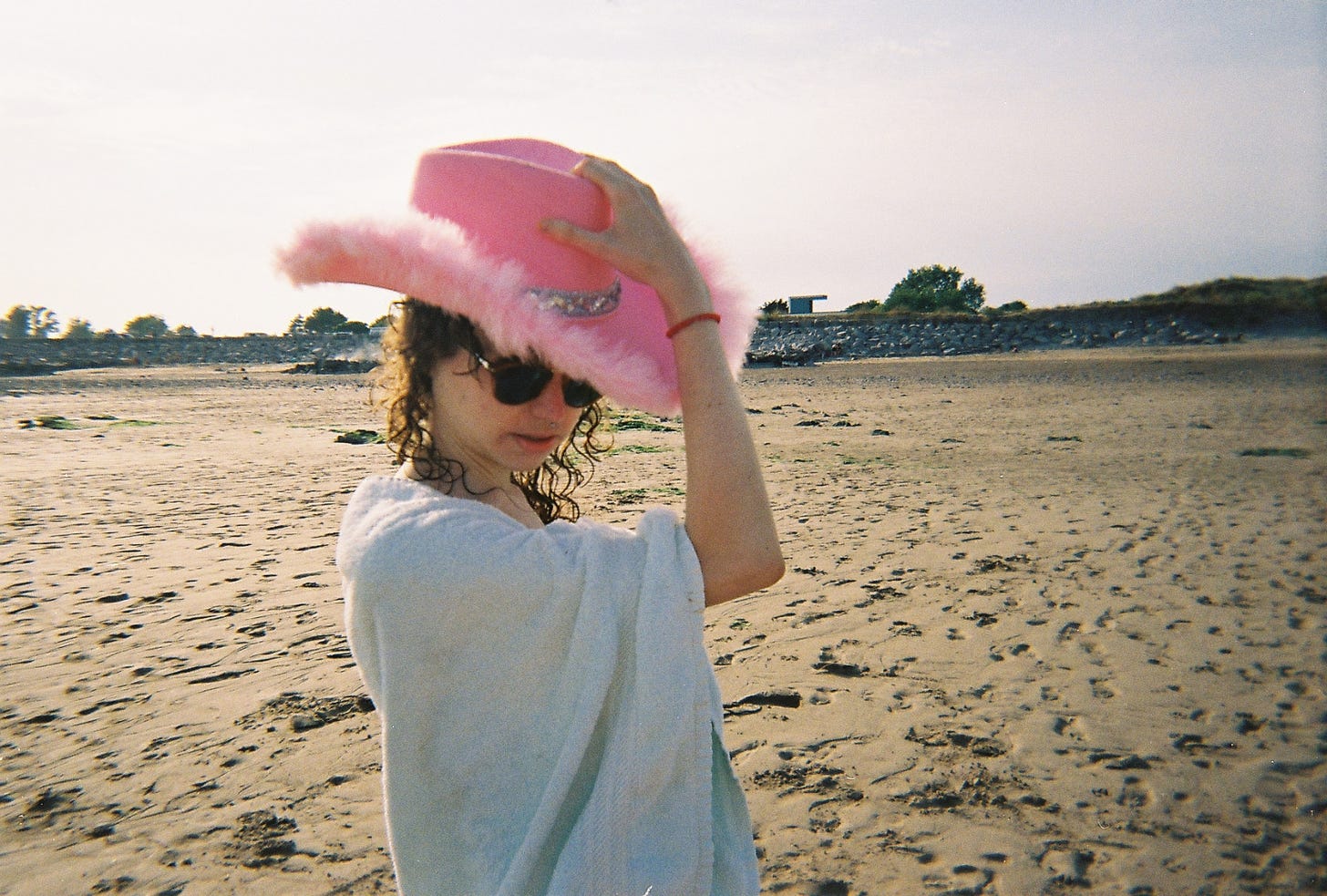 Photo of a person standing on a beach. They are a white femme presenting person, in the middle of the frame, wearing a pink cowboy hat and sunglasses, wrapped in a towel. Their hand is on the top of their hat like they are about to cock it in a sort of ‘howdy’ way. Behind them is sand and a line of trees on the horizon. 