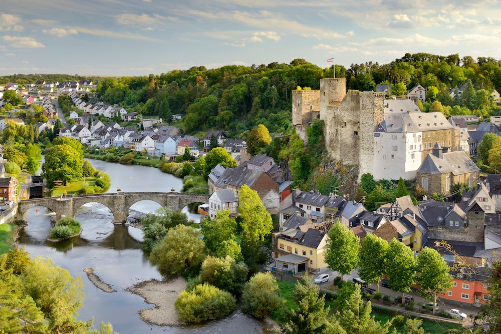 View of the River Lahn from high up, with a castle and medieval village to its right and an old stone bridge crossing over it. It is summer and the trees are bright green and the sky is blue and a little cloudy.