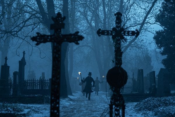 Figures holding torches hurry through a graveyard by twilight. A pair of crosses are closest to the camera.