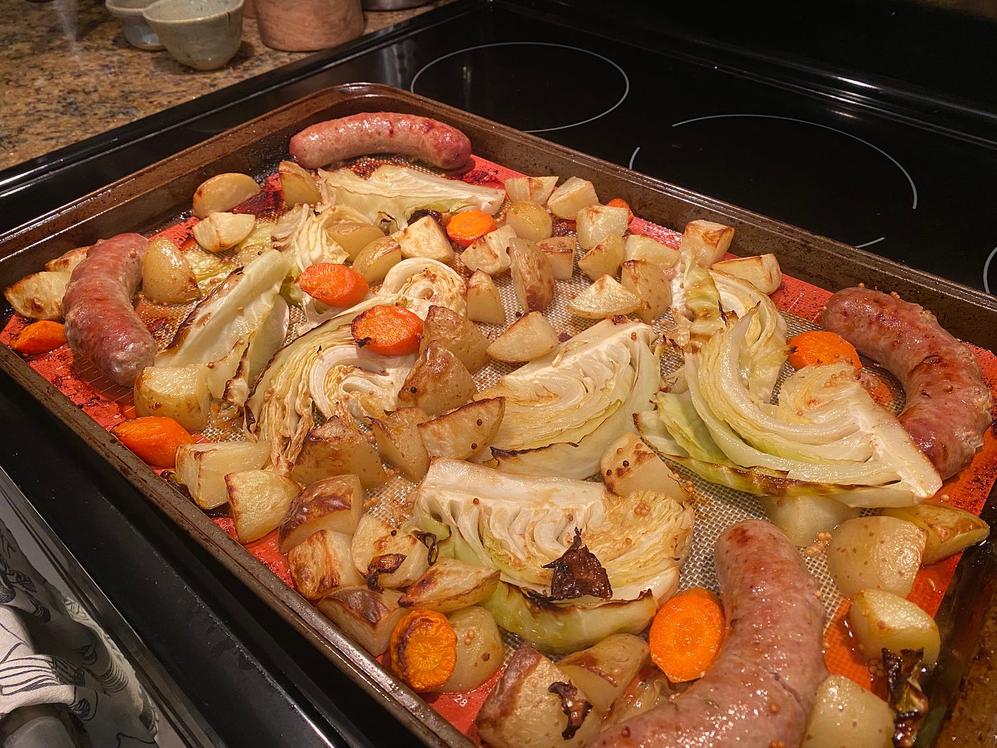 A large sheet pan lined with a silpat, covered with roasted cabbage, potatoes, and carrots, with four browned sausages at the corners.