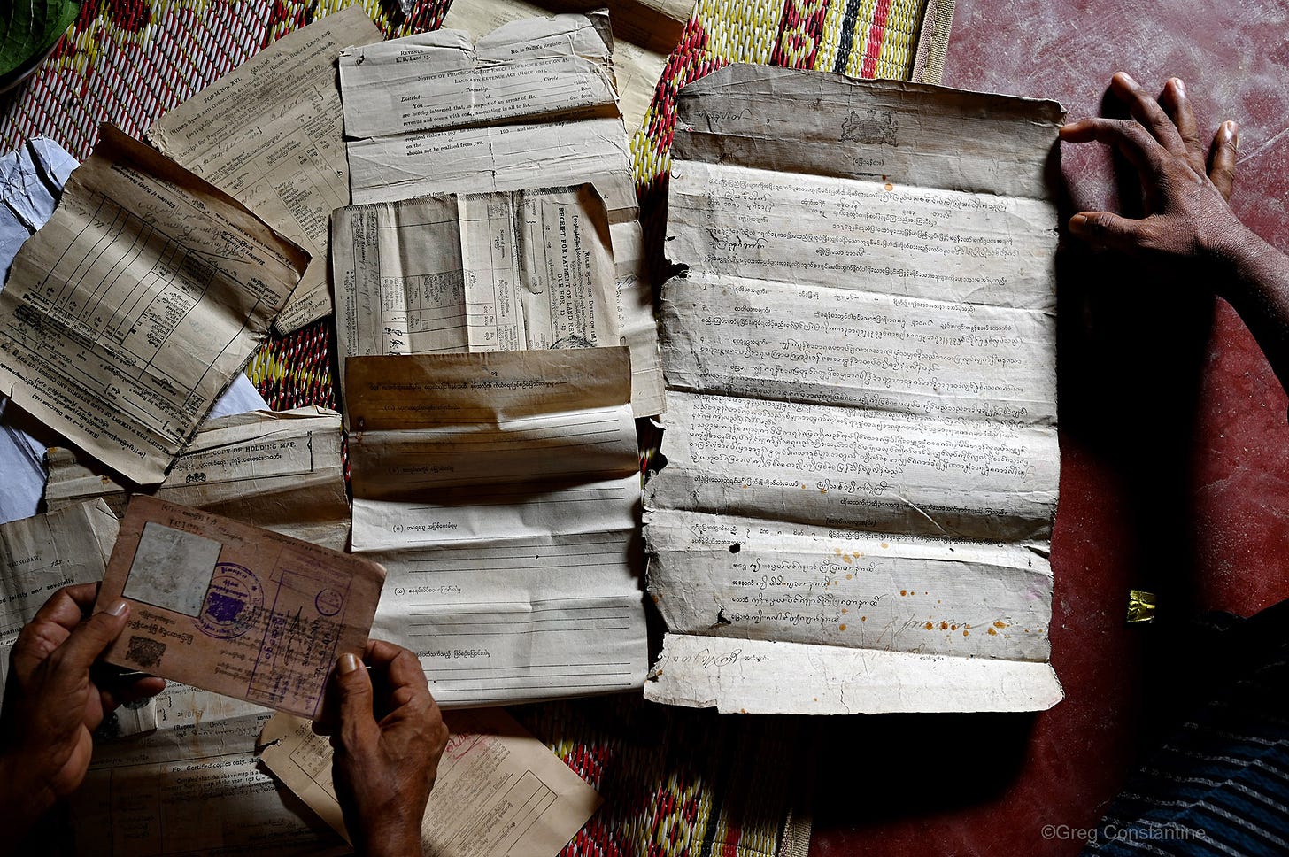 Picture of a handful of papers that are in the possession of a family of Rohingya refugees. The papers are spread out on a red floor and a colourful woven rug. In the top right a hand reaches for one paper; in the bottom left two hands are holding an envelope.