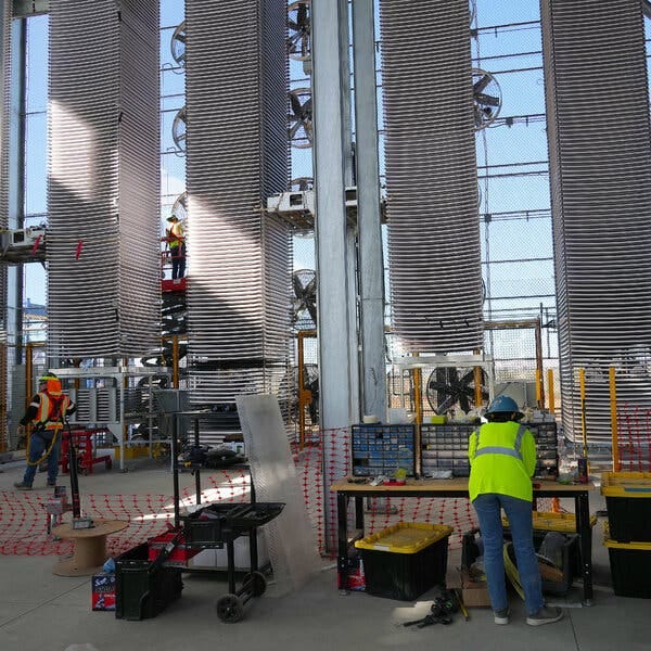 An open-air industrial site. In the foreground, a worker in a yellow vest and hard hat leans over a work bench. In the near background, tall stacks of trays. The sky beyond is clear blue. 