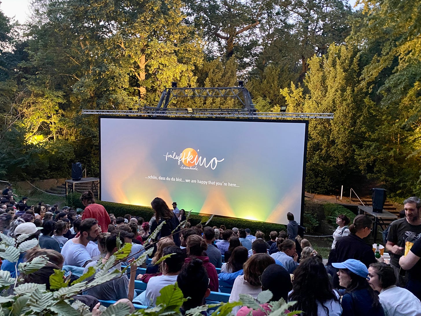 Imagem: Um cinema no meio de um parque, árvores atrás do telão e plateia lotada. Sol se pondo.