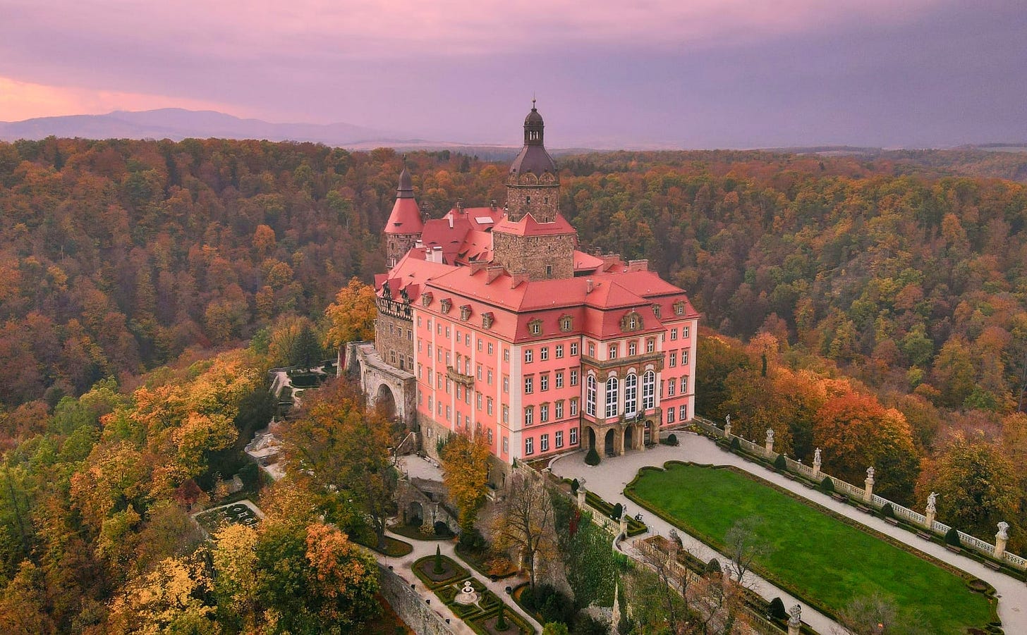 pink stone castle with a bell tower sitting among formal gardens on a hilltop surrounded by forest
