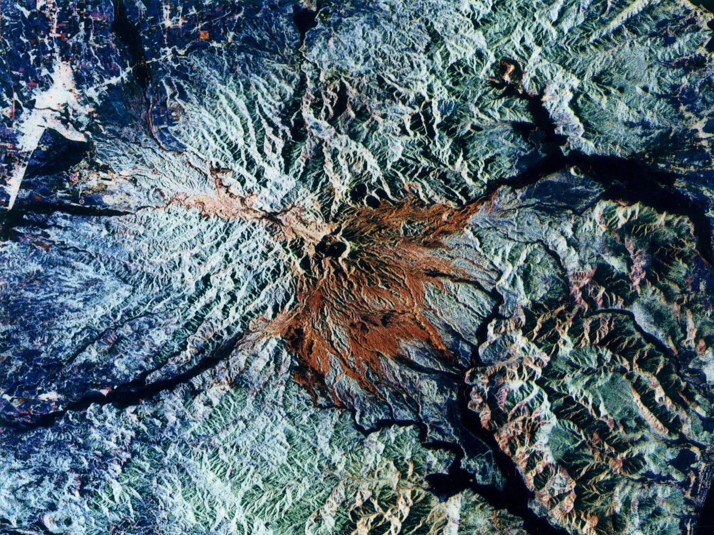 An aerial image of a volcano crater using Synthetic Aperture Radar, where the crater appears red and the surrounding moutains appear blue and green