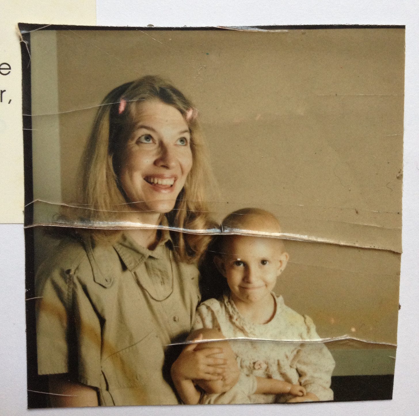 An old, wrinkled photograph in brown hues features Rebekah’s mother holding her  in her lap. Her mother looks up and to her left, smiling. Rebekah is bald from chemo treatments and looks straight toward the camera, her smile a tight line. She’s holding her baby doll, her hand over the doll’s eyes.