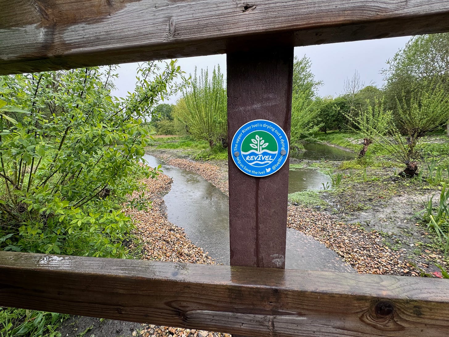 View through wooden fence looking out to the restored gravel beds and the start of the River Ivel chalk stream. On the central fence post is a RevIvel plaque which says, "The upper River Ivel is drying out and dying. Help us Save the Ivel (Twitter/X logo) @IvelRev"