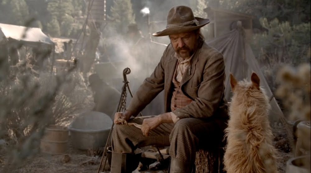 This image shows one of Deadwood's best characters, gold prospector Whitney Ellsworth (played by Jim Beaver) sitting in his prospecting camp near Deadwood. A dog, with its back to camera, sits at Ellsworth's feet.