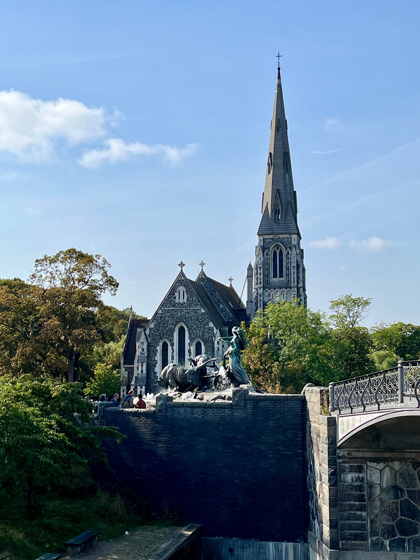 gray stone building with a tower and spire beside it, a bridge in front and a fountain statue of a woman driving bulls before her. Blue Sky and trees surrounding the building, water running below the bridge.