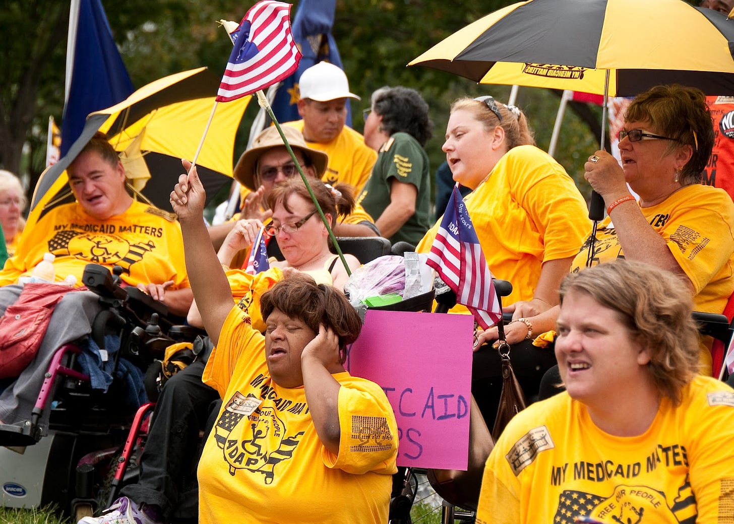 A crowd of ADAPT disability activists wearing yellow shirts are waving signs and flags in support of Medicaid access A crowd of ADAPT disability activists wearing yellow shirts are waving signs and flags in support of Medicaid access