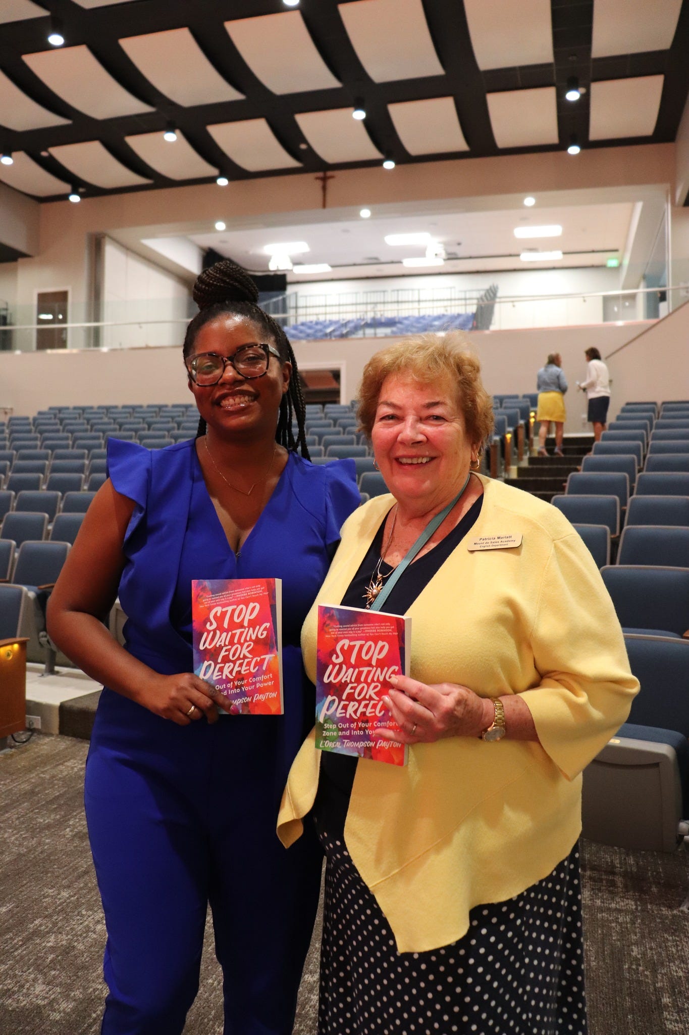 Picture of a Black woman wearing a blue jumpsuit standing next to a white woman wearing a yellow cardigan. Both are holding books in their hands.