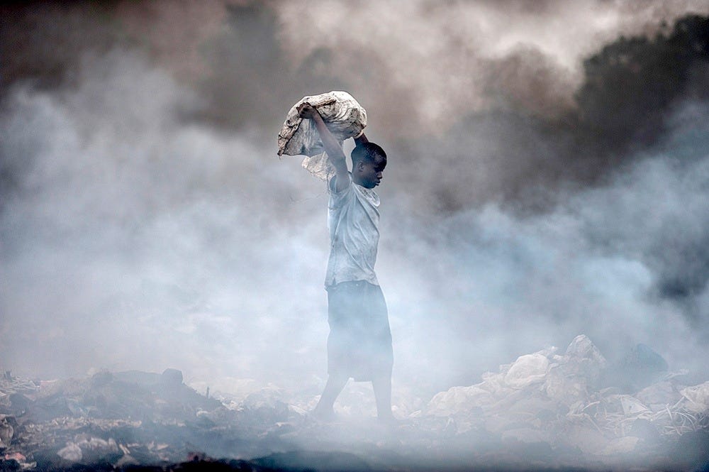 A black boy, maybe 12 or 13, hoists a bag above his sholders as he crosses a smoldrring dump.