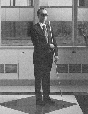 A black-and-white photo of a man with dark, short hair. He wears sunglasses, a suit, and a tie. He holds a cane. and wears dark shoes.