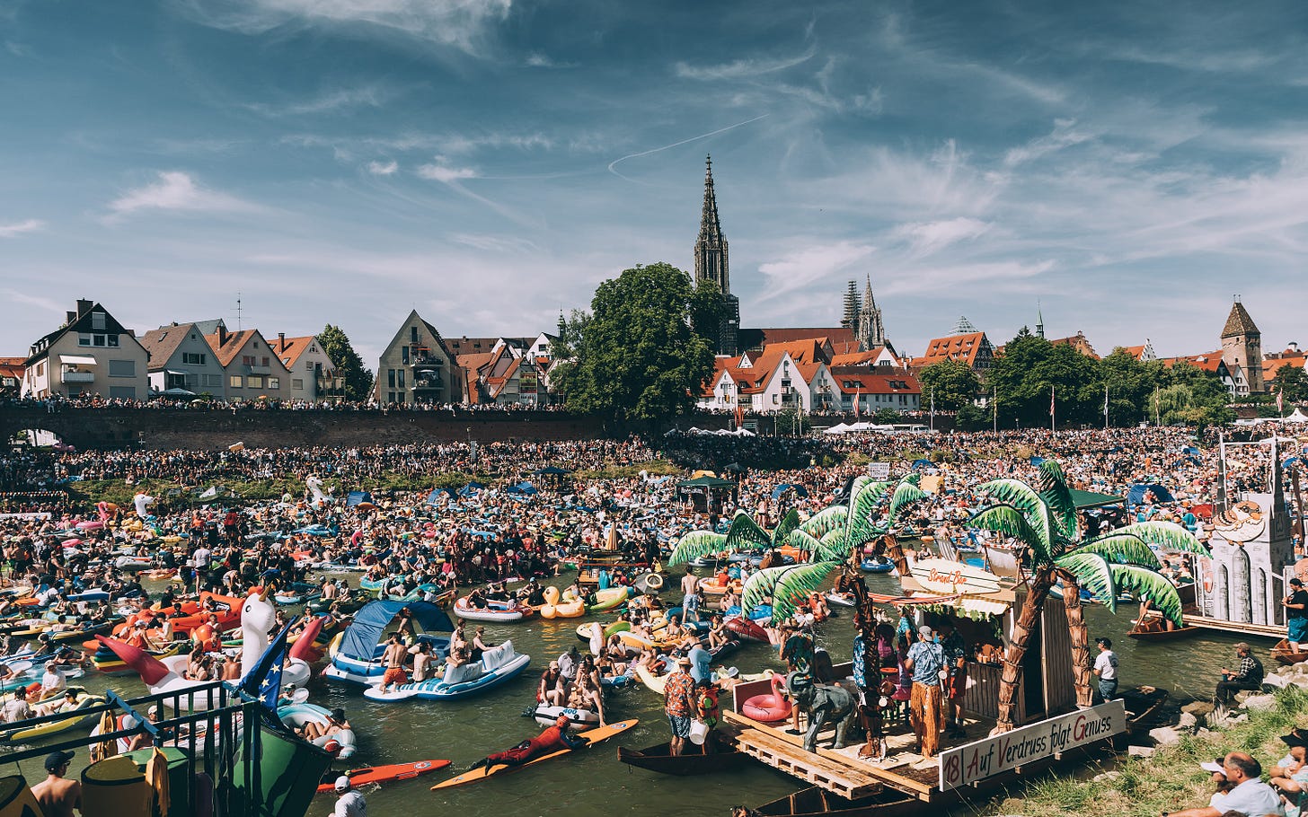 A chaotic scene of a river filled with people and inflatables, with floats that have palm trees on. There are also paddle boarders. In the background there are buildings and onlookers.