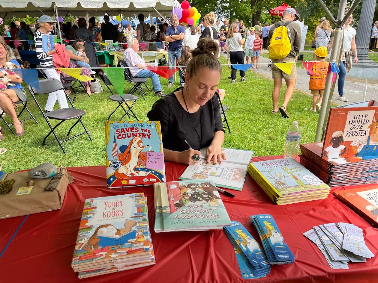 The author, a white lady with her hair pulled into a bun, sits at a table signing a picture book she wrote. I am writing this in the third person, but it's me, Julie Falatko, I am the lady. There are other books and bookmarks and people and dogs and balloons in the background.