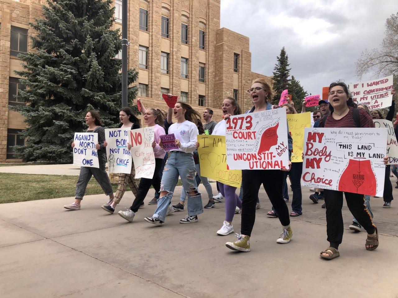 On a gray, cloudy day, protesters, mainly young women march in the foreground bearing pro-abortion rights signs, in front of the sandstone buildings of the University of Wyoming campus.