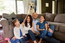 A family sitting on a couch with a dog in the background

Description automatically generated