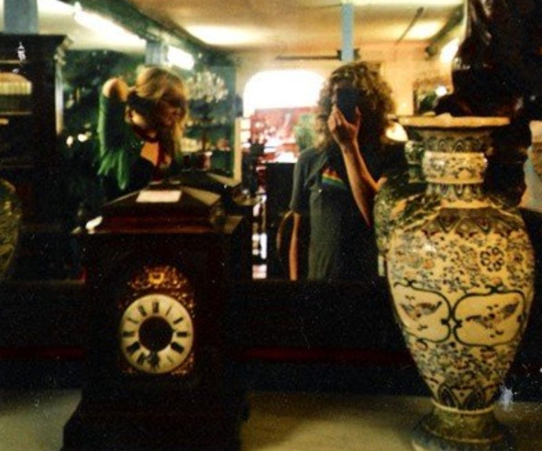 Lizzy and Jez, two eighteen year olds, reflected in a mirror in an antique shop, between a carriage clock and a large vase. 