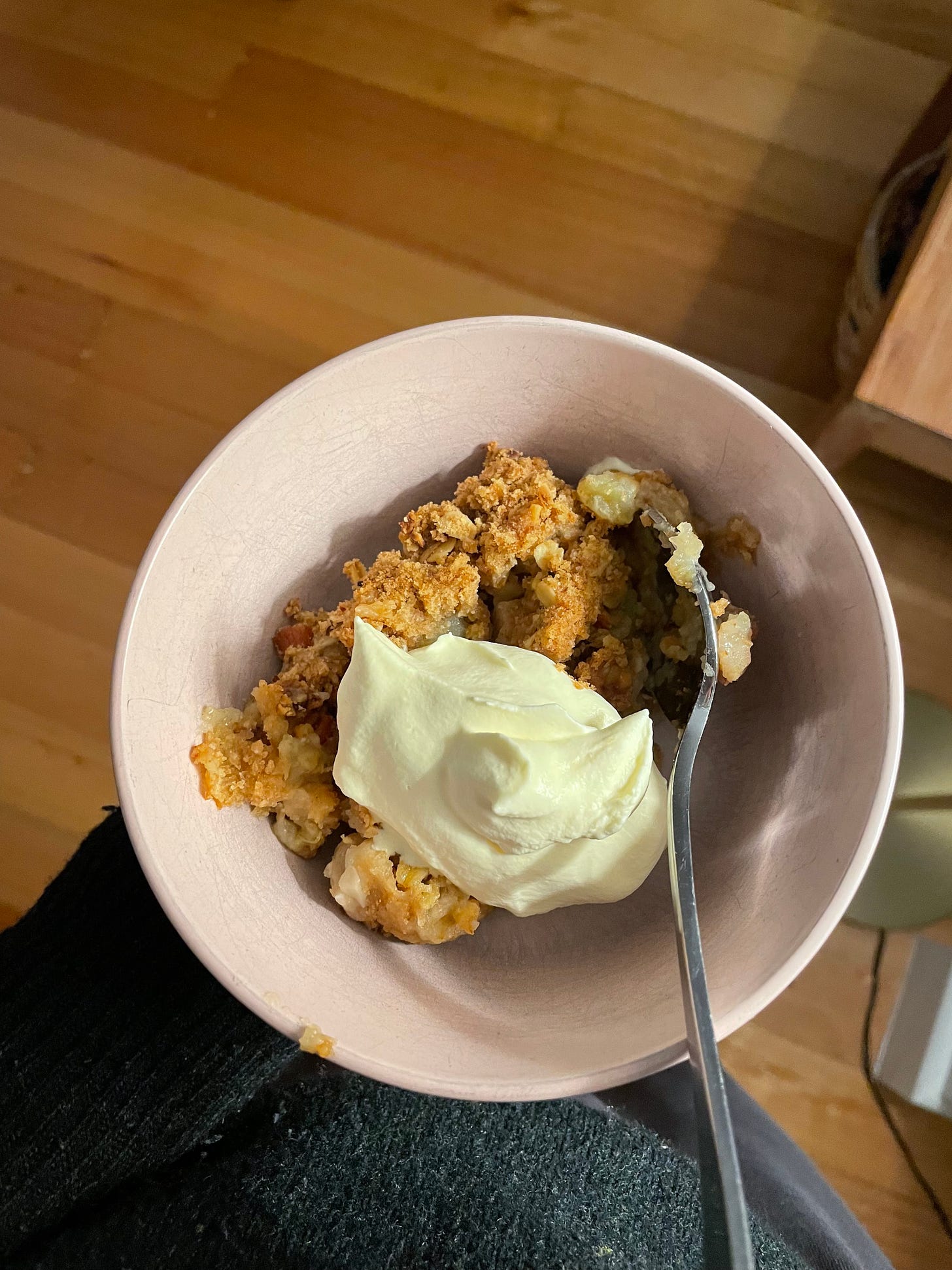 Small bowl of apple crumble, with a golden-brown topping. Served with whipped cream on top. The person holding the bowl is wearing a dark green woollen jumper and track pants for a night in at home.