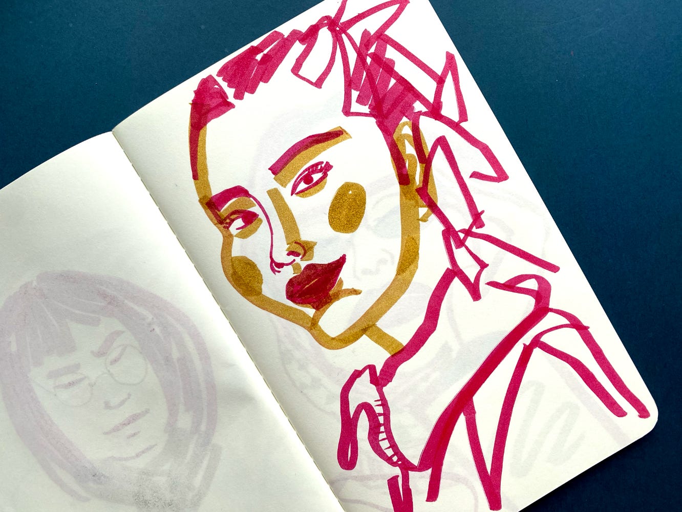 2 colour drawing of a female face in a open sketchbook laid on a flat dark blue surface