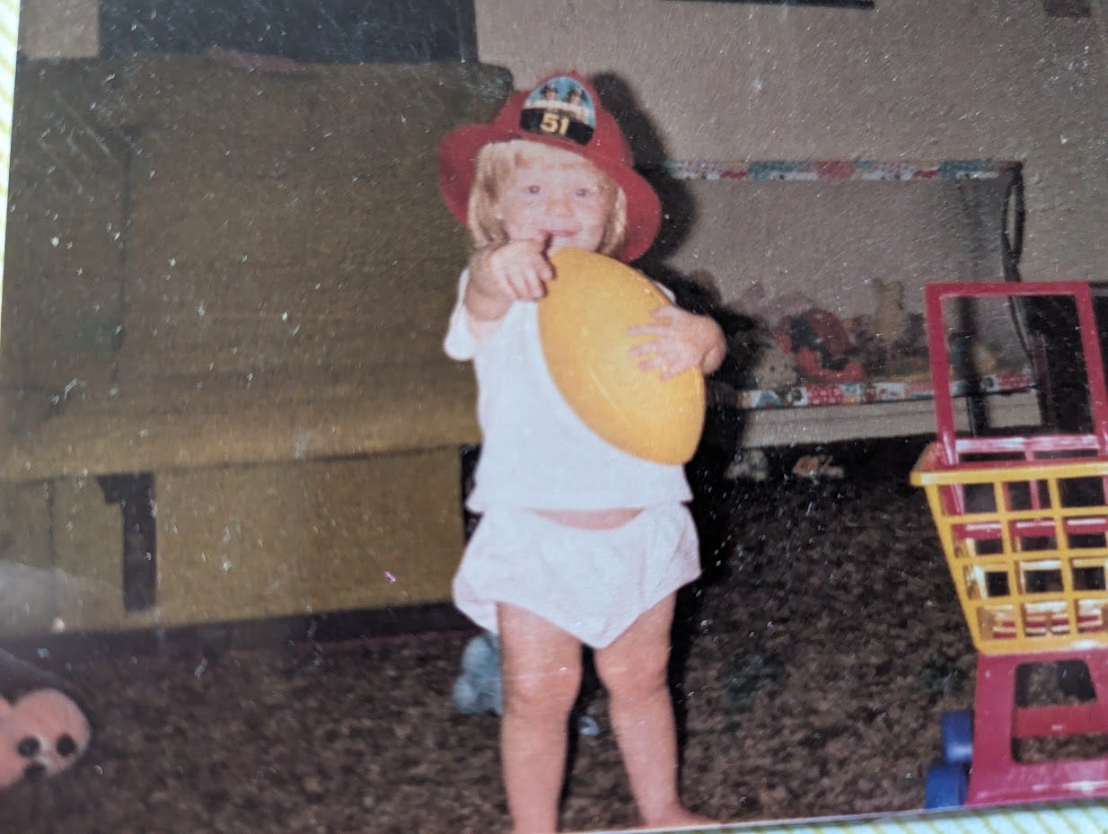 A young child wears a red firefighter hat, holds a yellow ball, and points to the camera, toys and a couch in the background