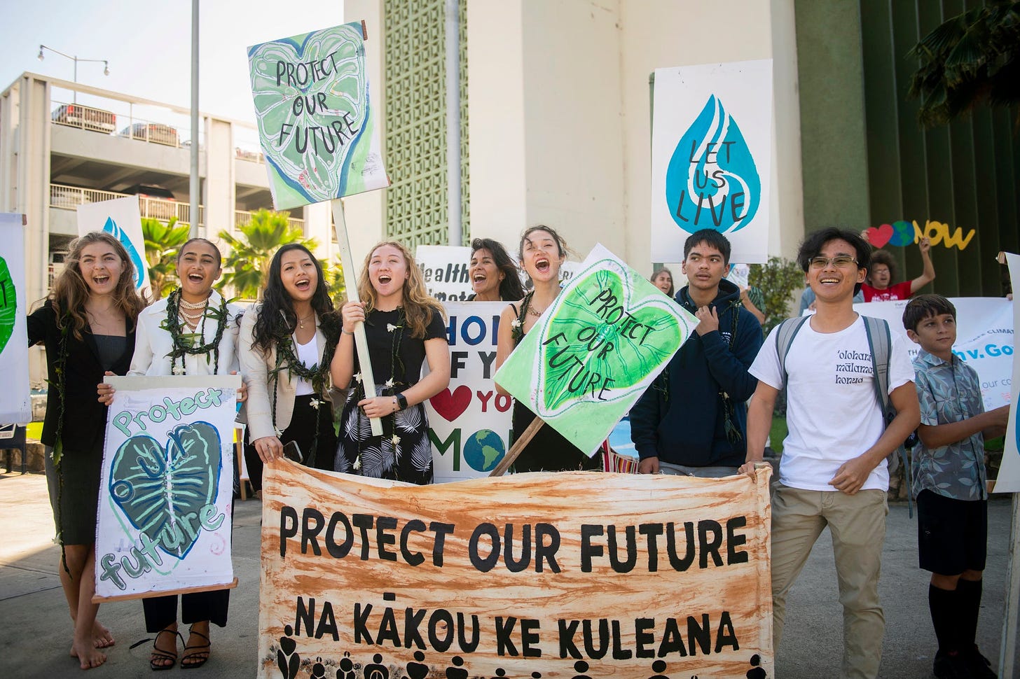Eight teens are upbeat and excited during rallySeven or eight teenagers on a sidewalk outside an urban building holding signs with heart-shaped kalo leaves that read Protect Our Future and Let Us Live
