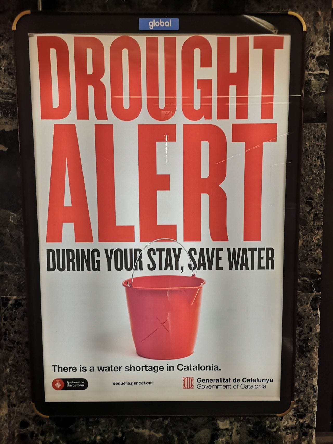 Poster with "drought alert" written on it, showing an empty bucket and asking people to save water while staying in Catalunya. 