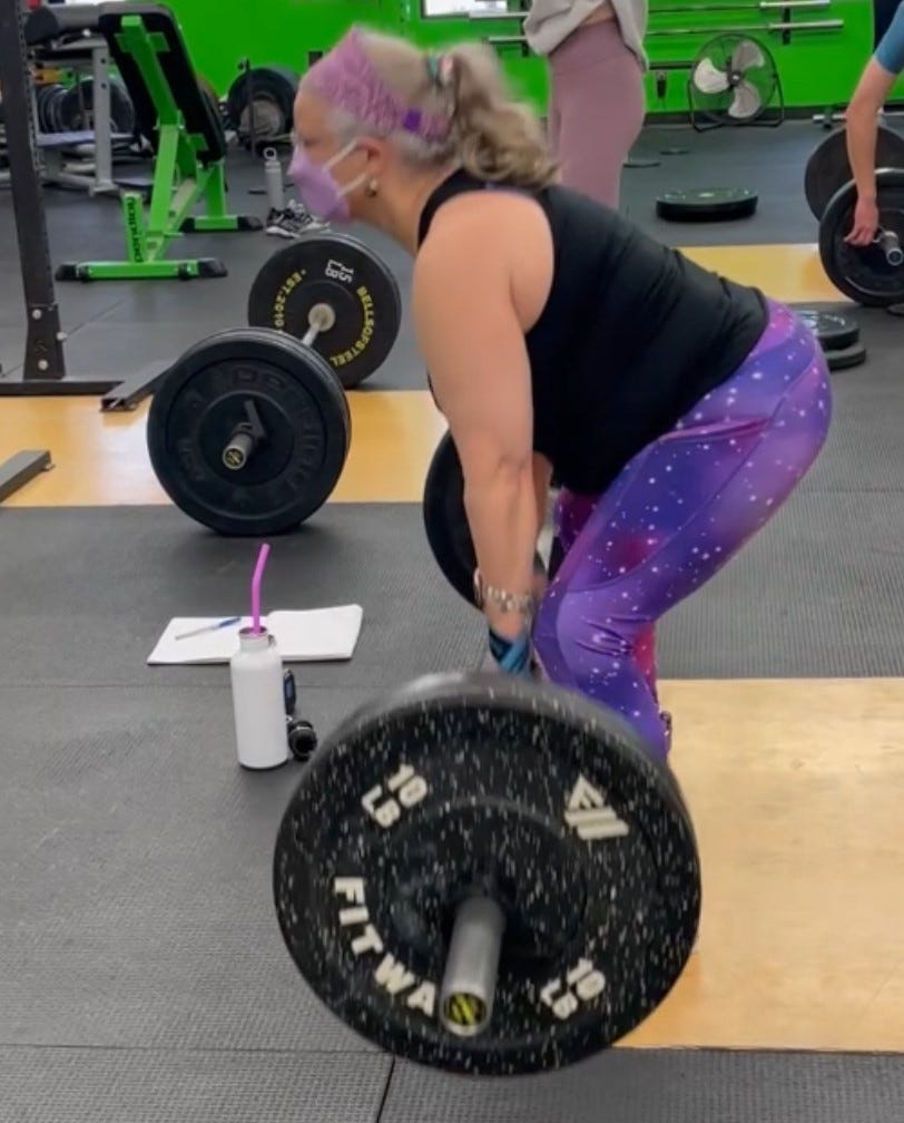 silver haired femme in a black top and purple leggings, headband and mask, doing some deadlifts