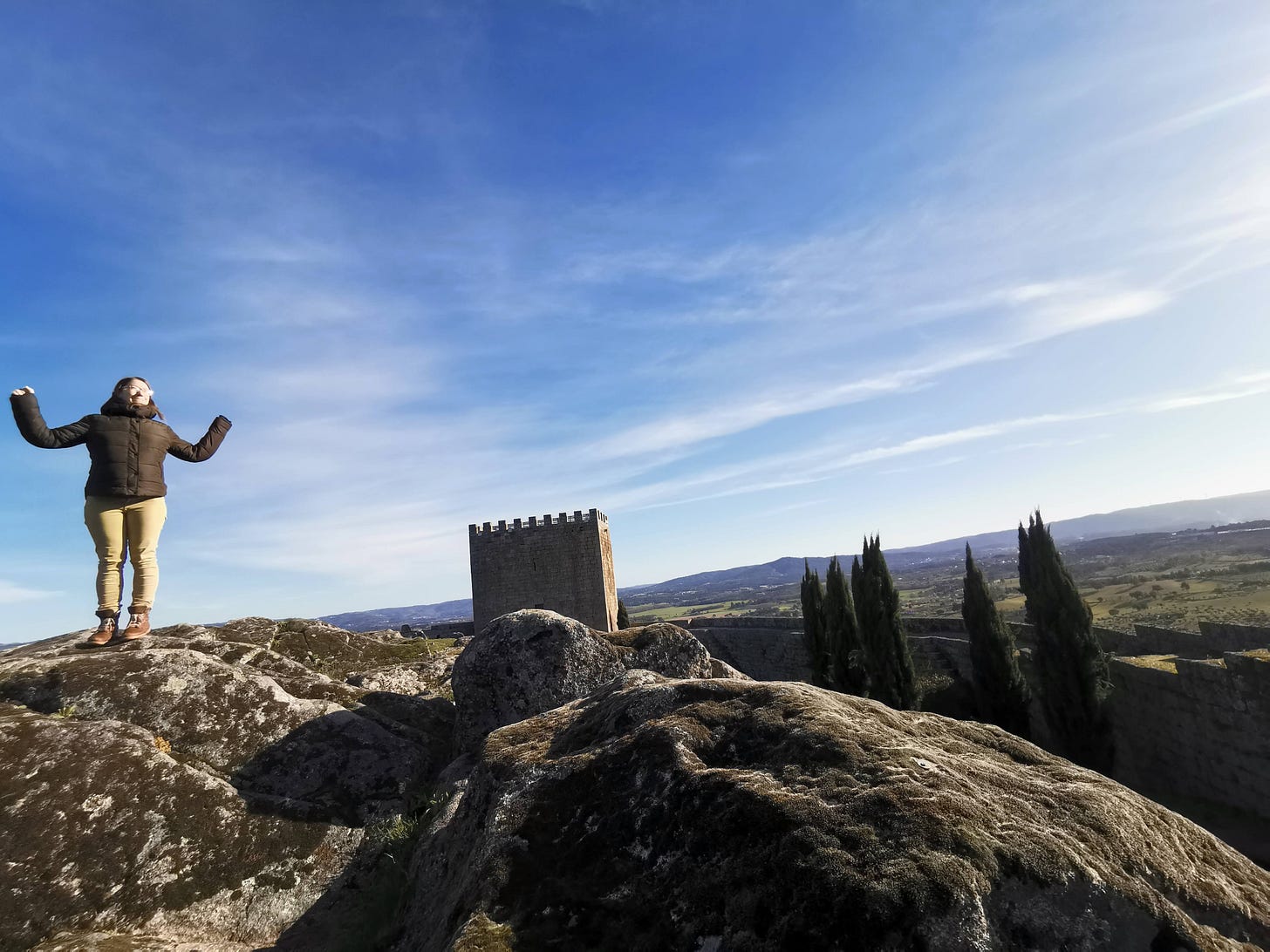 A woman standing on top of rocks with her arms raised to the sky in a vast landscape on top of a hill with a castle wall and trees in the background