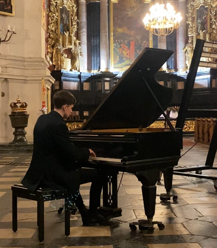 A pianist in a large, fancy building. The room is ornate, with gold trims and classical paintings. The pianist is dressed in a suit, and there is a crown in the background.