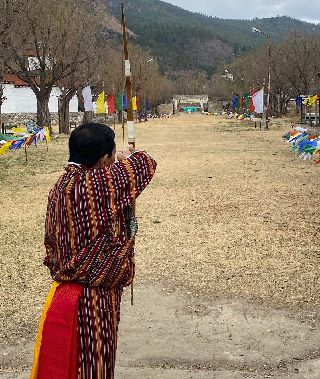 An archer, dressed in a traditional gho takes aim at a target approximately 150 feet away. Along path to the target are colorful prayer flags