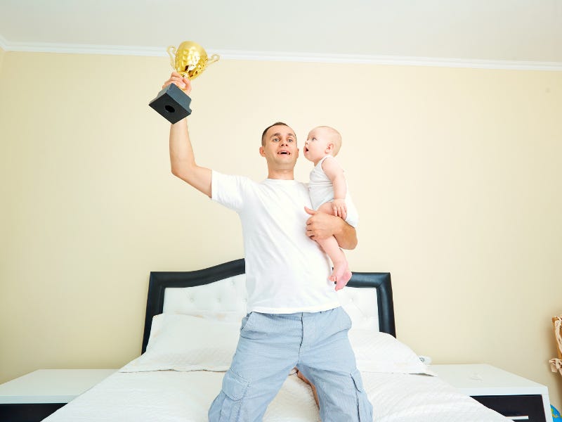 A bloke kneeling on a bed, holding a baby in one arm, whilst holding aloft a gold trophy with the other.