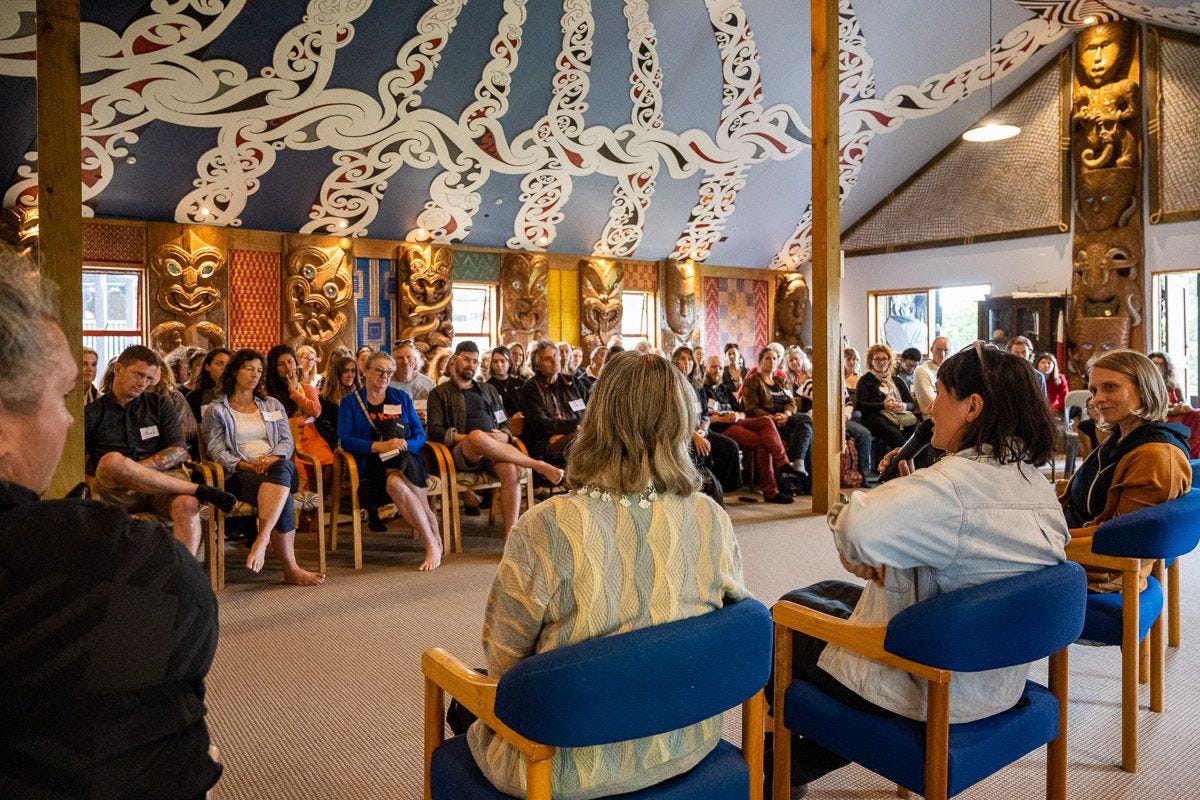 People sitting in chairs in a Wharenui, facing an audience listening to a talk about food systems. The carvings of the Wharenui are visible in wood at the back and in painted panels on the ceilings.