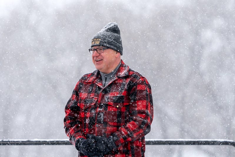 Minnesota Gov. Tim Walz smiles during a snowstorm, wearing a redplaid jacket, ski cap and gloves