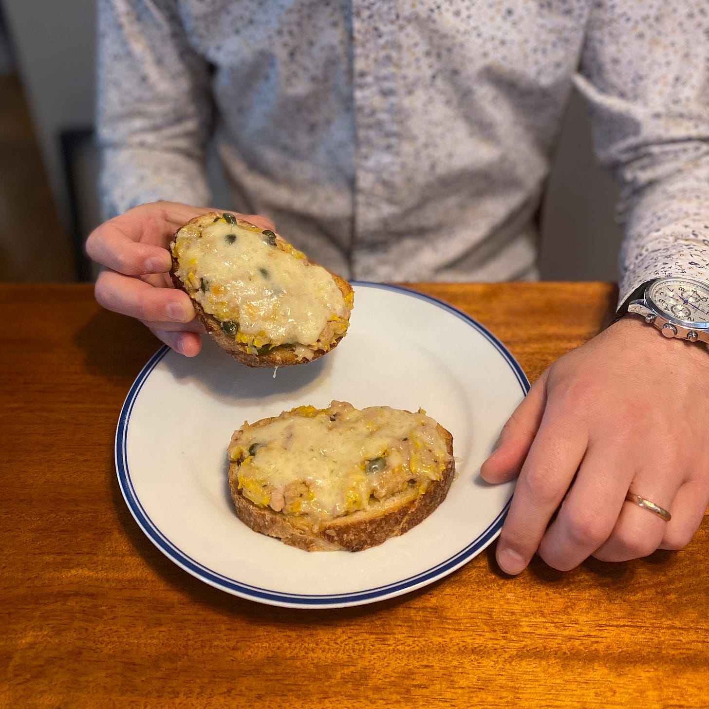 A white and blue side plate with two slices of sourdough, each with the tuna melt described above. One of Jeff's hands rest next to the plate, and the other is holding one of the slices of toast.