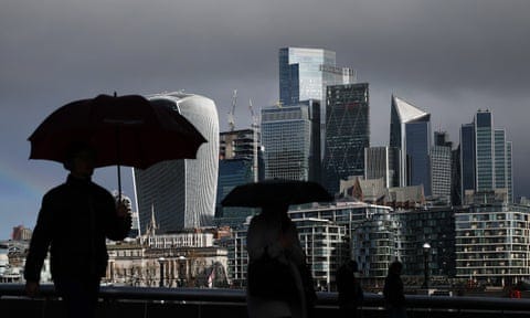 Commuters in silhouette cross London Bridge with umbrellas up, near the City of London's skyscrapers