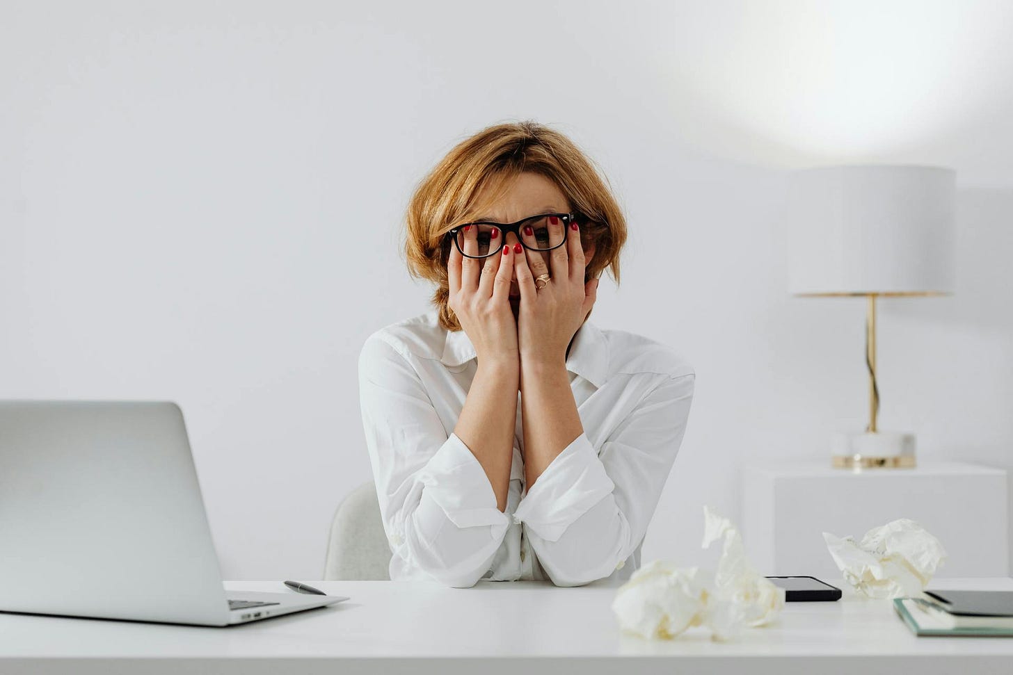 Woman in White Long Sleeve Shirt Wearing Eyeglasses Sitting at a Table holding her face