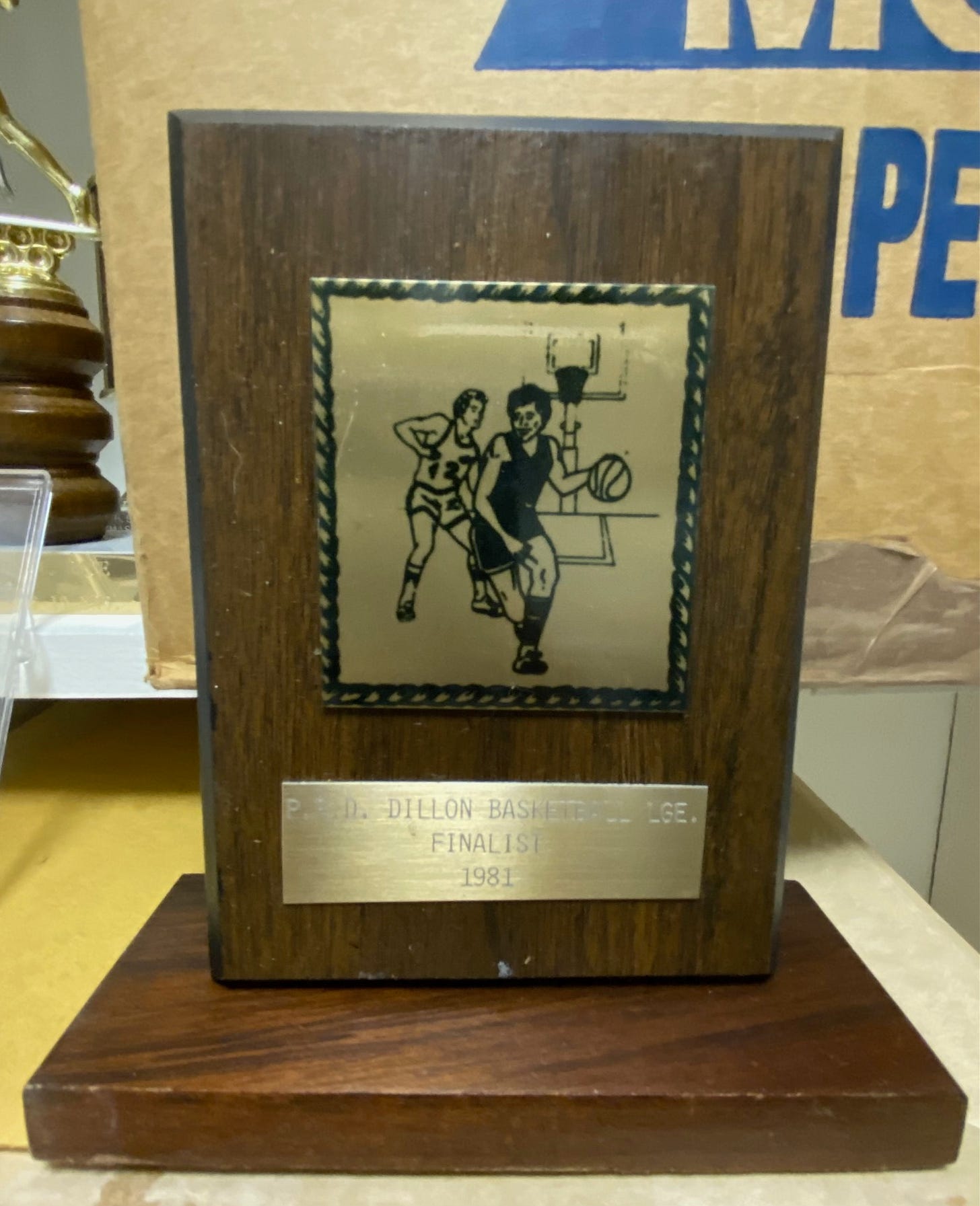 Dark brown wooden plaque, showing a male player dribbling past another player. Inscription reads: Dillon Basketball Lge. Finalist 1981. Dark brown wooden plaque, showing a male player dribbling past another player. Inscription reads: Dillon Basketball Lge. Finalist 1981.