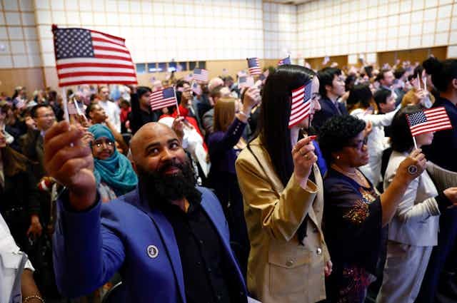 A large crowd of people waving small American flags are crowded into a small room. In the front left corner, a man wearing a purple jacket smiles.