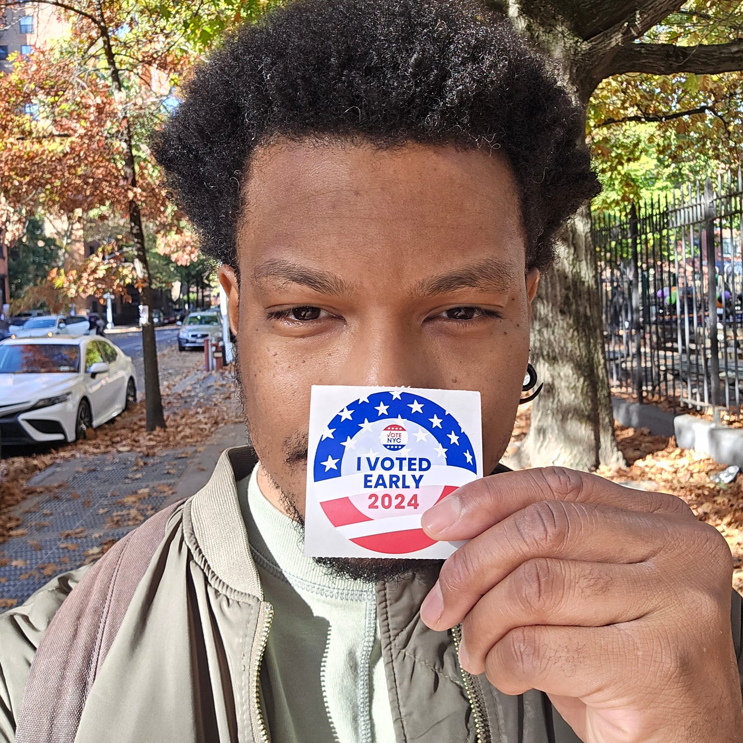 A close-up self-portrait of the author, a young Black man with natural hair, wearing an army green jacket, holding up an "I VOTED EARLY 2024" sticker from NYC. The photo is taken outdoors on a tree-lined street during fall, with autumn leaves visible in the background.