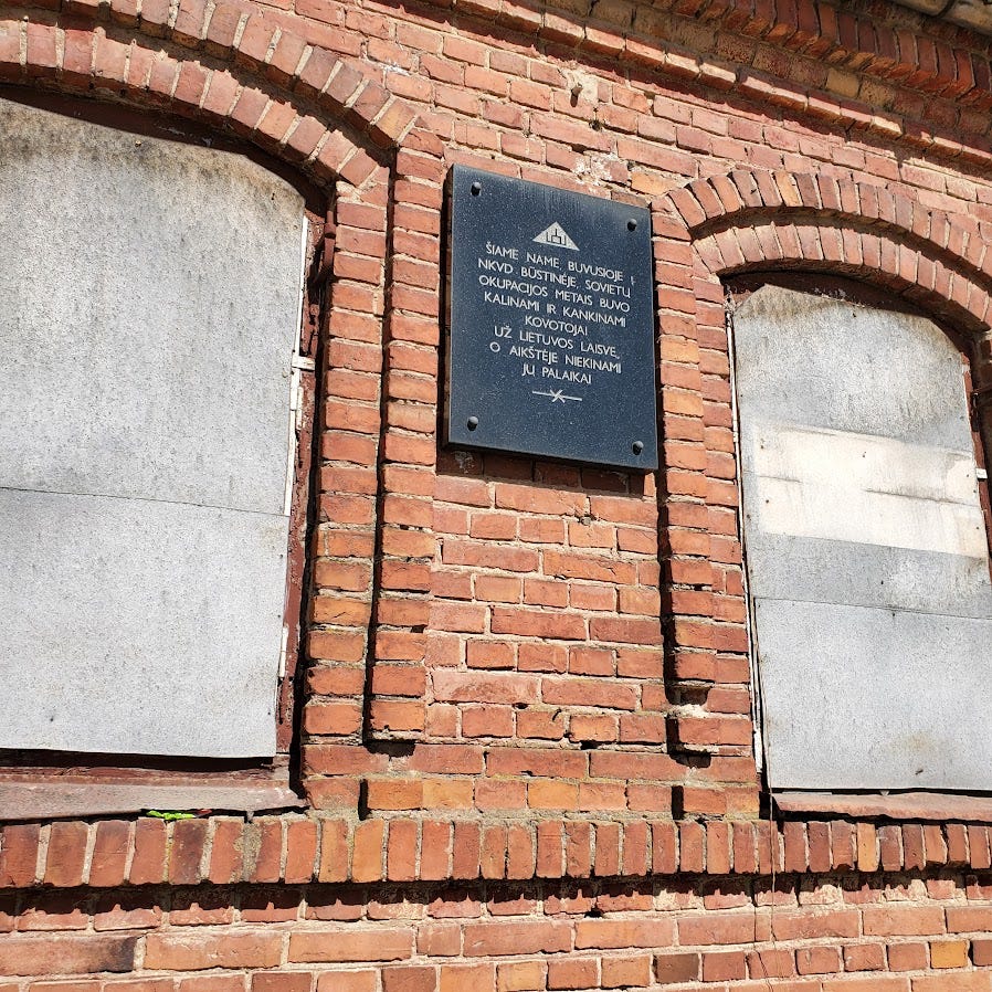 A black plaque with writing in Lithuanian on a brick wall surrounded by two boarded up windows.