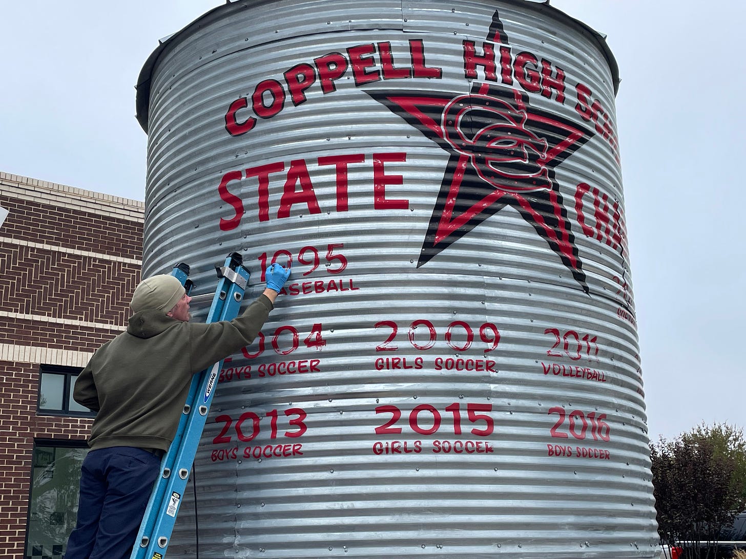 A man on a ladder touches up the paint on a silo listing Coppell High School's state championships