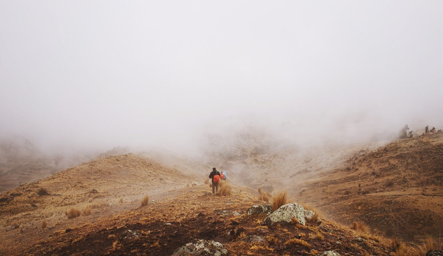 Imagem: um vale montanhoso com neblina e três pessoas de costas caminhando muito distante