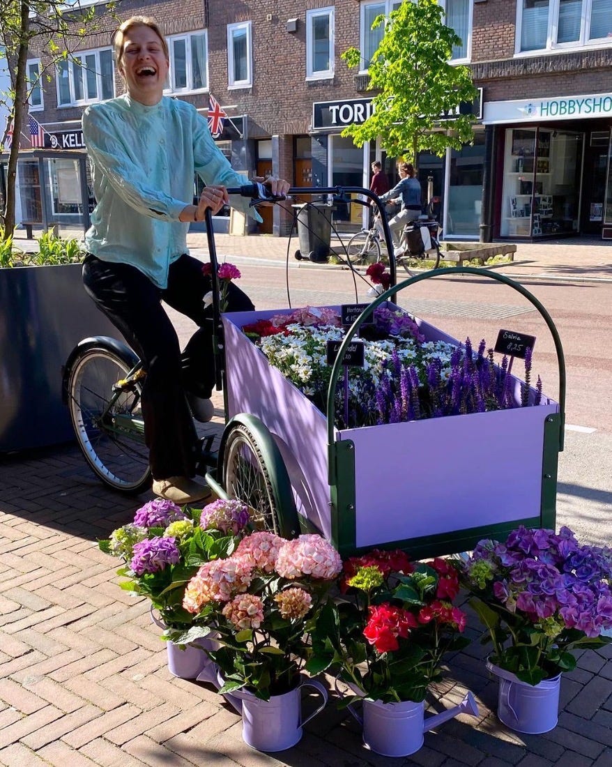 A woman, presumably the owner of the florist, sits on the cargobike and laughs. The cargobike is parked on the pavement and not travelling. The box is full of flowers for sale. In front of the cargobike are other potted flowers for sale. 