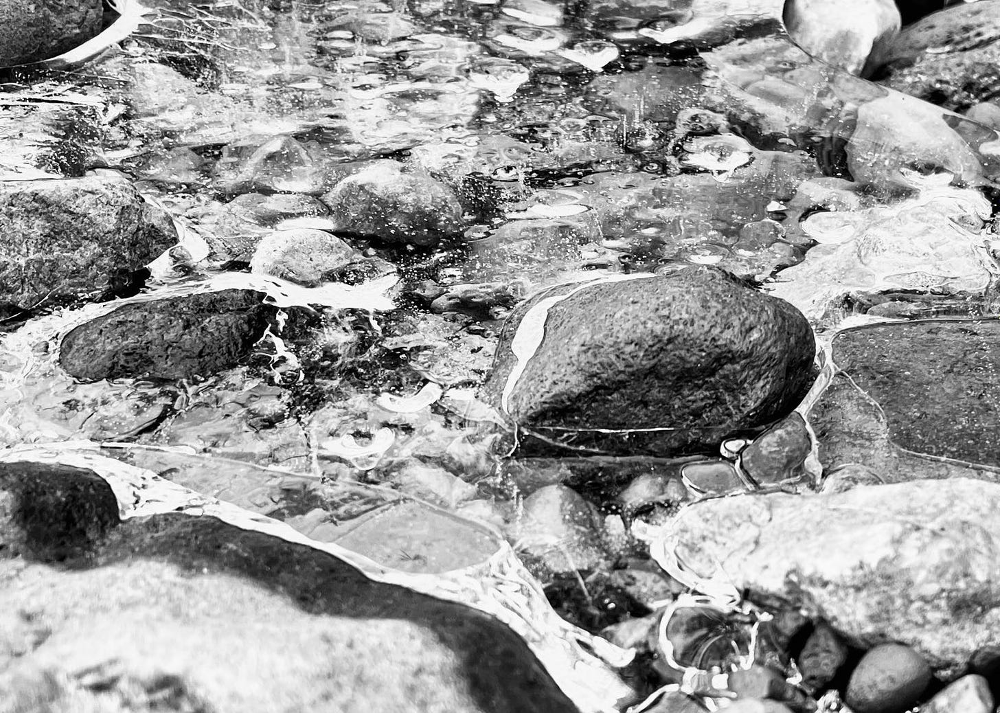 A black and white photo of a frozen stream bed. Several of the larger rocks are partially submerged in the ice. The ice is mostly clear with some cracks and bubbles visible throughout.