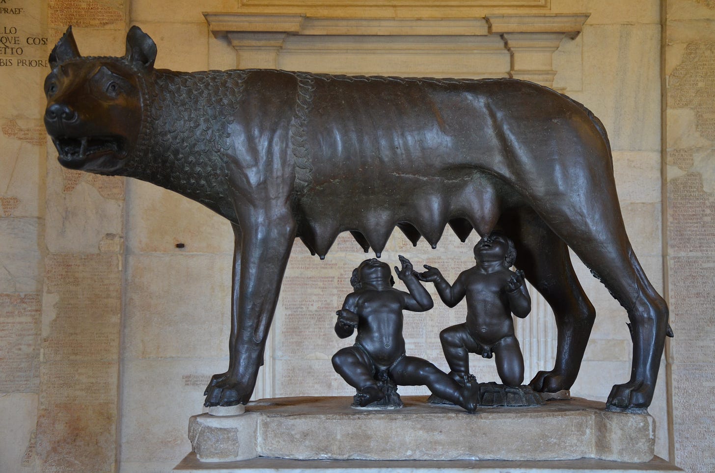 The Capitoline Wolf, a bronze wolf with mouth agape, looking off into the distance with its mouth open, while Romulus and Remus (two cherubic toddlers) stand below the wolf's breasts, awaiting milk