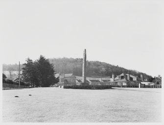 Black and white photo of Sueno's Stone, a tall carved cross slab, standing on grassy parkland with houses behind and a wooded hill behind that.