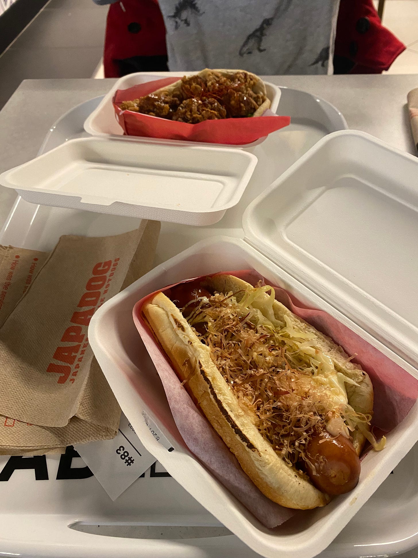 On a white tray are two white paper takeout containers, each with their own Japadog. In the foreground is mine, with bonito flakes and green cabbage. Steph's, with chicken karaage, is in the background. Brown napkins scattered on the tray read 'JAPADOG' in red block letters.