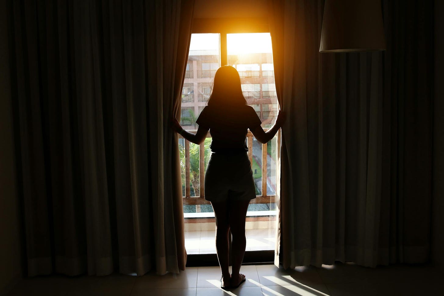 Photo of a woman in dark room standing in front of a window with the curtains cracked open to reveal a sunny day outside