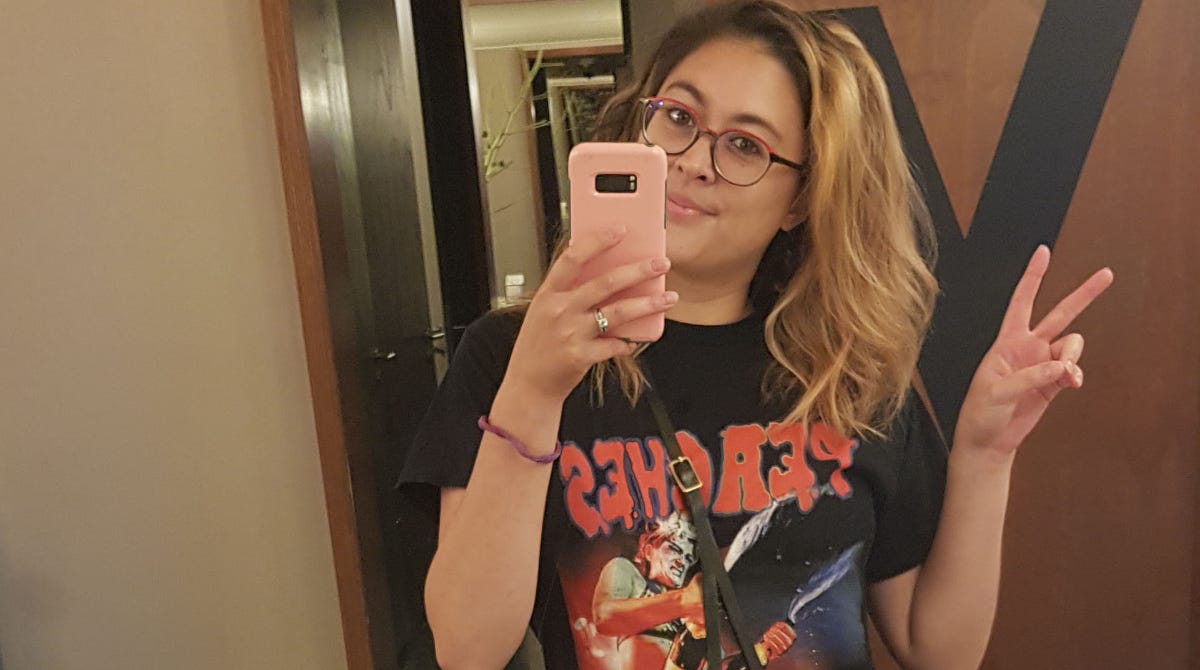 woman with bleached hair and round glasses takes a picture of herself with a phone. The black t-shirt reads "Peaches" but backwards. She holds her index and middle fingers in the shape of a V.